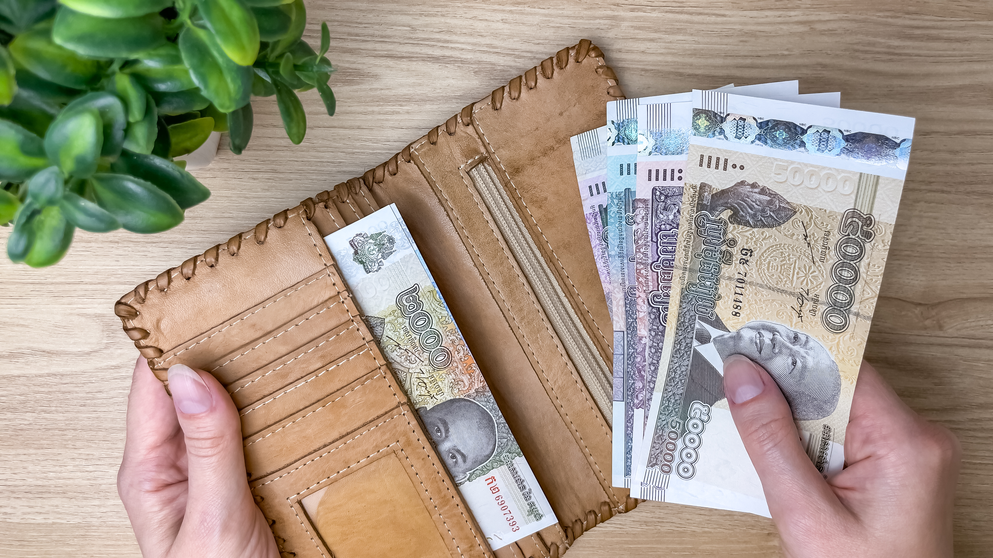 A woman's hands take Cambodian riels from a leather wallet on a wooden table. The national currency of Cambodia, a concept for personal finance, cash spending, and budgeting.